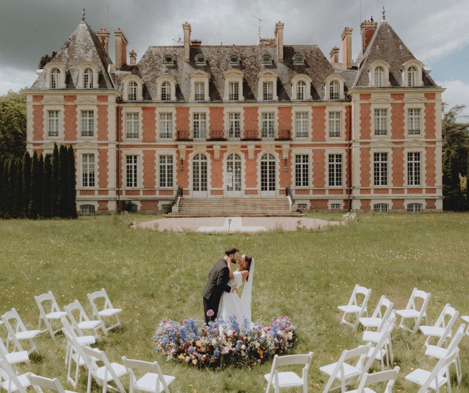 Pretty bright floral arrangement on the lawn of the Chateau de la Cazine with the historic chateau in the background.Bride and Groom leaning in for a kiss infront of a circular arranged ceremony space of white chairs.