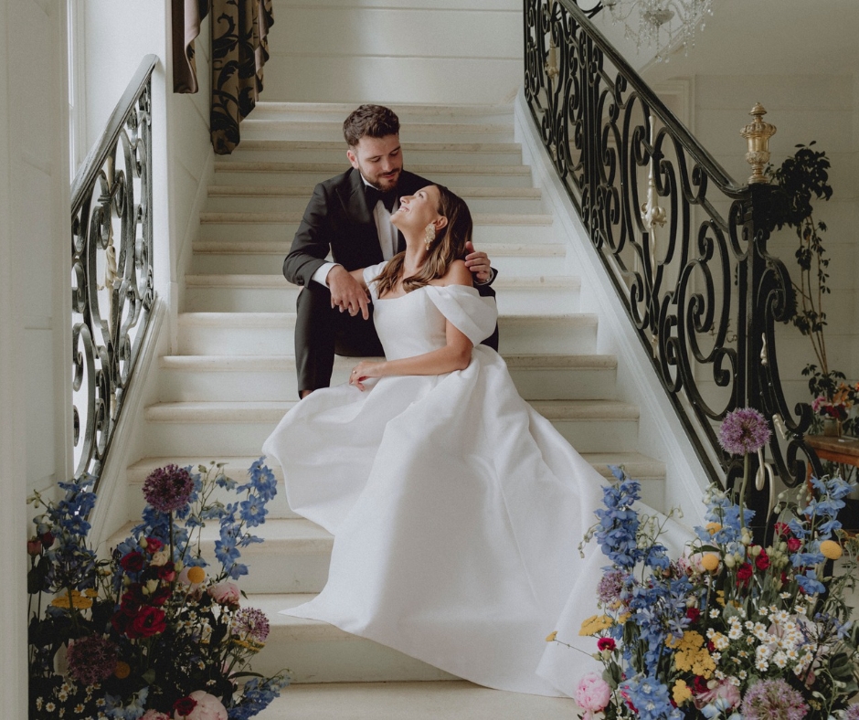 Bride and Groom sit on the ornate black staircase at the Chateau de la Cazine adorned with colourful florals- daisies peonies and Violets