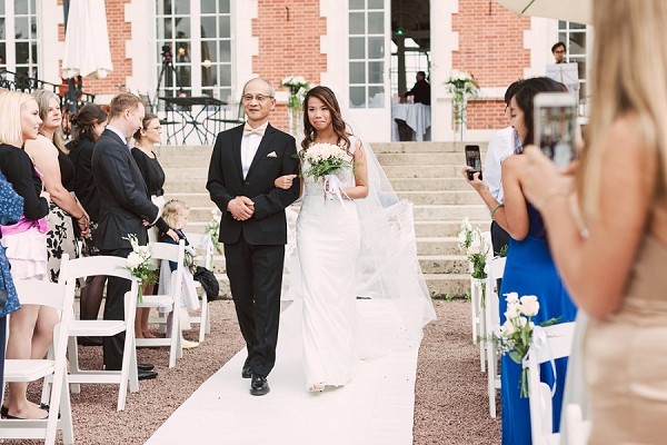 Father walking the bride down the aisle at an outdoor ceremony at Chateau de la Cazine with the red brick building as the backdrop